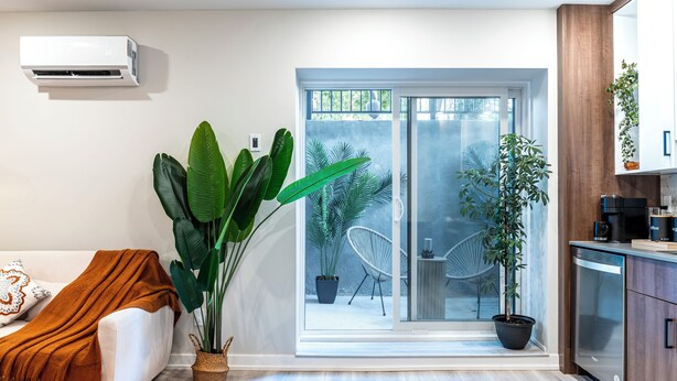 A modern living room with a white sofa, a large green plant, and a view of a patio with outdoor furniture.