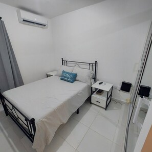 Quarto individual com uma cama de solteiro, colcha branca, mesa de cabeceira com luminária, gaveta pequena e ar condicionado fixado na parede.