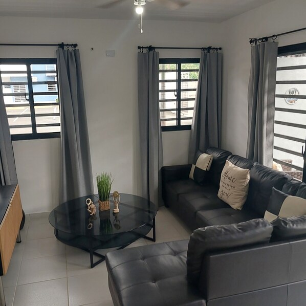 Uma sala de estar moderna com um sofá de couro preto, uma mesa de centro de vidro e cortinas cinzas.