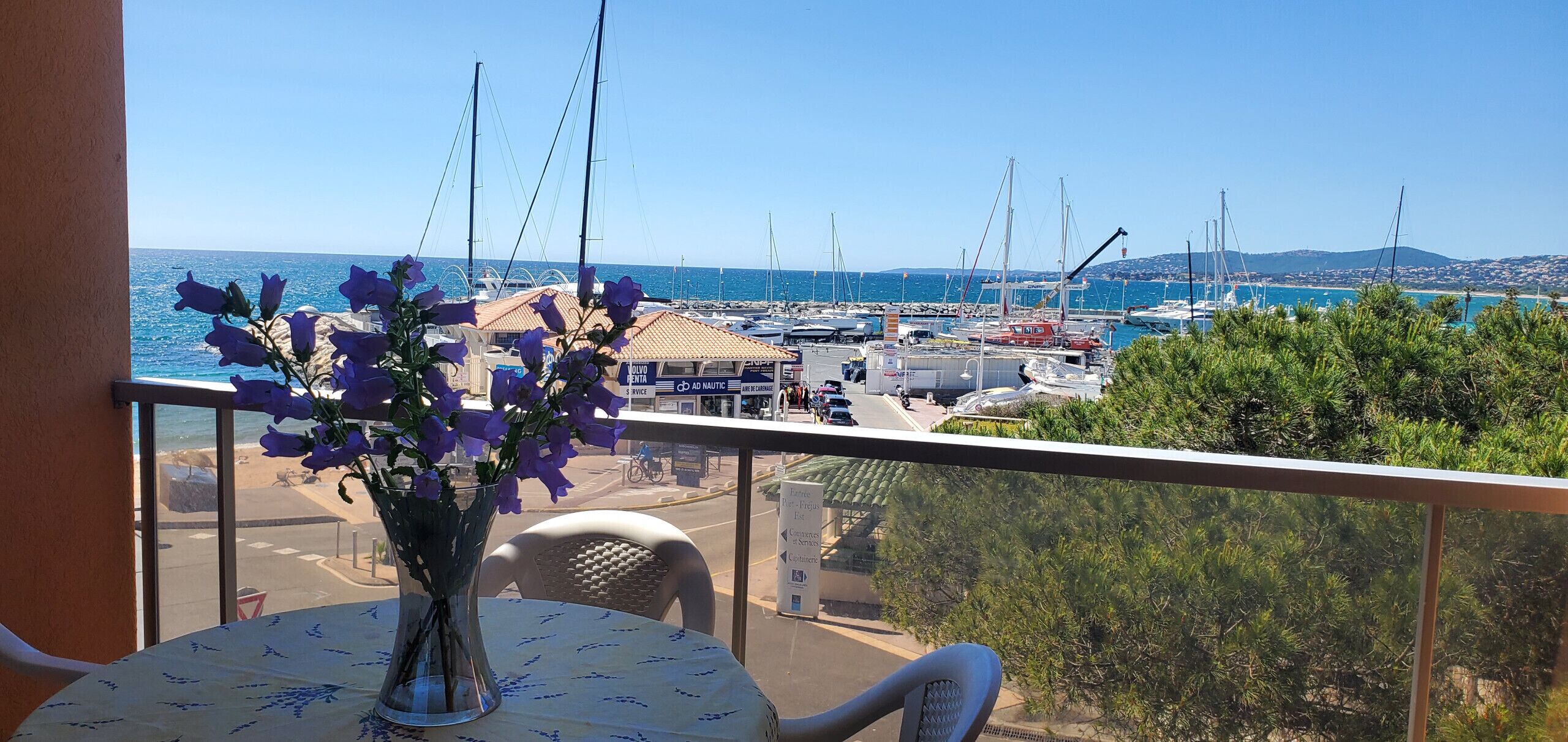 A balcony with a table and a vase of flowers, overlooking a marina with boats and a clear blue sky.