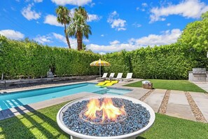 A backyard with a swimming pool, a fire pit, lounge chairs, and a palm tree.