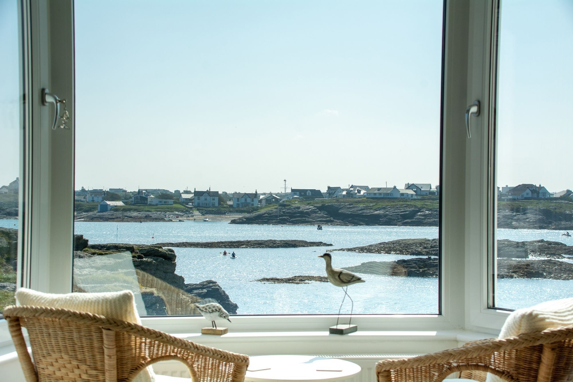 Vista desde una habitación con ventanas amplias que dan a la costa, con casas y terreno rocoso.