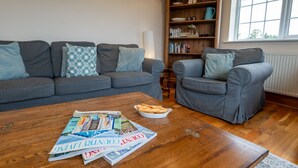 Un salon avec un canapé gris, un fauteuil gris et une table basse sur laquelle on trouve des magazines et un bol de gressins.