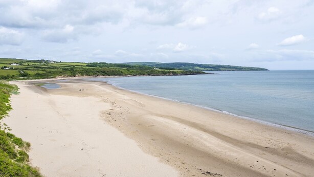 Une vaste plage de sable fin aux vagues douces, une colline verdoyante et, au loin, une autre colline parsemée de bâtiments.