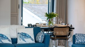 A dining area with a table set for two, a blue sofa with patterned cushions, and a window with a view of the outside. - 113 Cae Du, Abersoch (Abersoch)