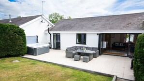 A patio with a hot tub, a grey sectional sofa, and a glass table. - 113 Cae Du, Abersoch (Abersoch)