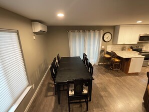 A dining area with a long table and chairs, a kitchen with white cabinets and a clock, and a window with blinds. - Comfy 3-bed bungalow in East Wenatchee, where nearby adventures await. (East Wenatchee)