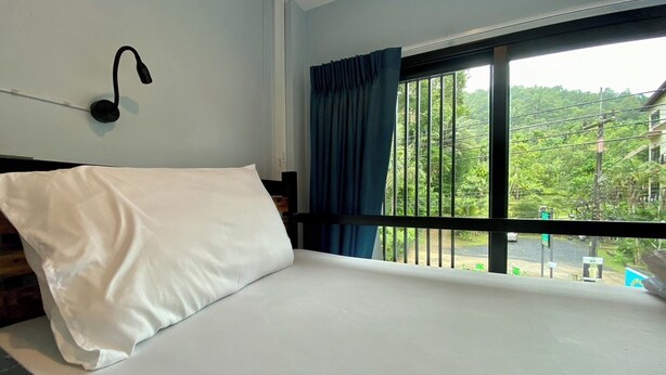 A bedroom with a bed, a window with curtains, and a wall-mounted light fixture. - Boom Hostel Khao Sok (Phanom)