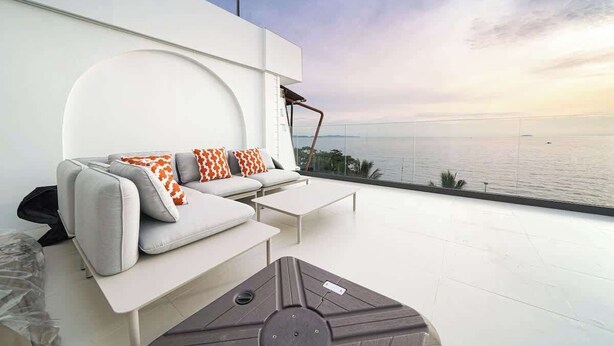 Ein moderner Außenbereich mit einer Couch und einem Couchtisch, einer Lampe und Blick aufs Meer.