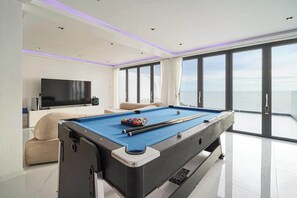 Una mesa de billar en una habitación moderna con un ventanal grande, un televisor de pantalla plana y un sofá.