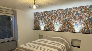 A bedroom with a bed, a window with blinds, a wall-mounted light fixture, and a floral-patterned wallpaper. - Appartement la Citadelle (Belfort)