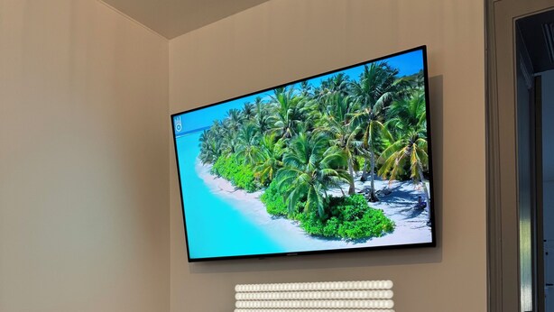 A modern living room with a flat-screen TV mounted on the wall displaying a tropical beach scene, a wooden console table with a framed picture and a vase, and a wooden floor. - Appartement la Citadelle (Belfort)