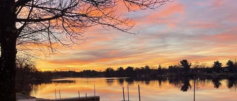 En rolig sø ved solnedgang med fortøjede både og et træ i forgrunden.