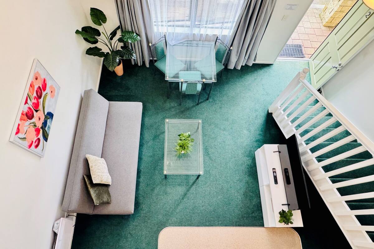 A modern living room with a grey sofa, a glass coffee table with a plant, a dining table with chairs, and a staircase with white railings.