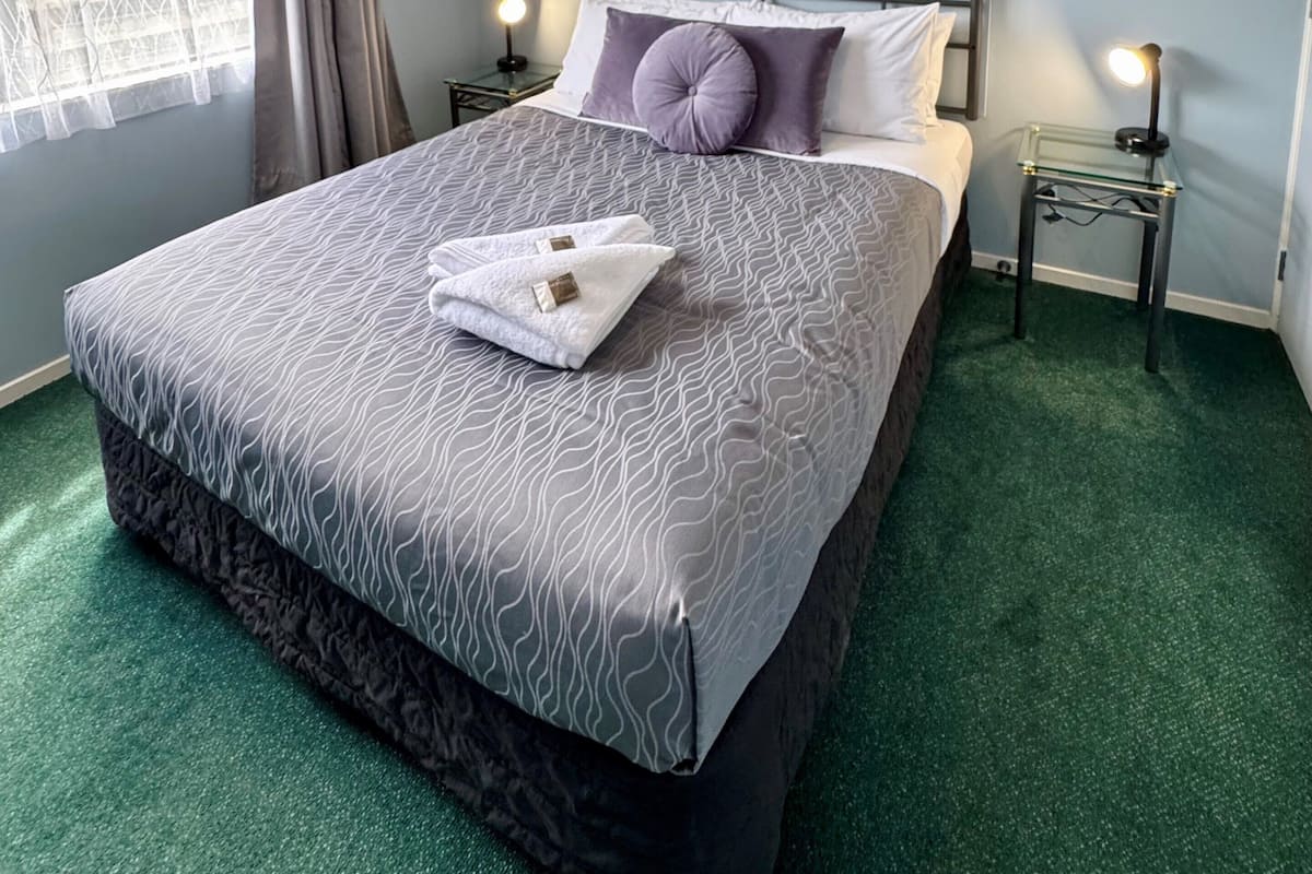 A neatly made bed with a patterned bedspread, two pillows, and a folded towel on a green carpeted floor.