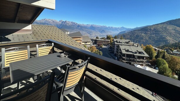 Ein Balkon mit Tisch und Stühlen mit Blick auf eine gebirgige Landschaft mit Gebäuden und blauem Himmel.