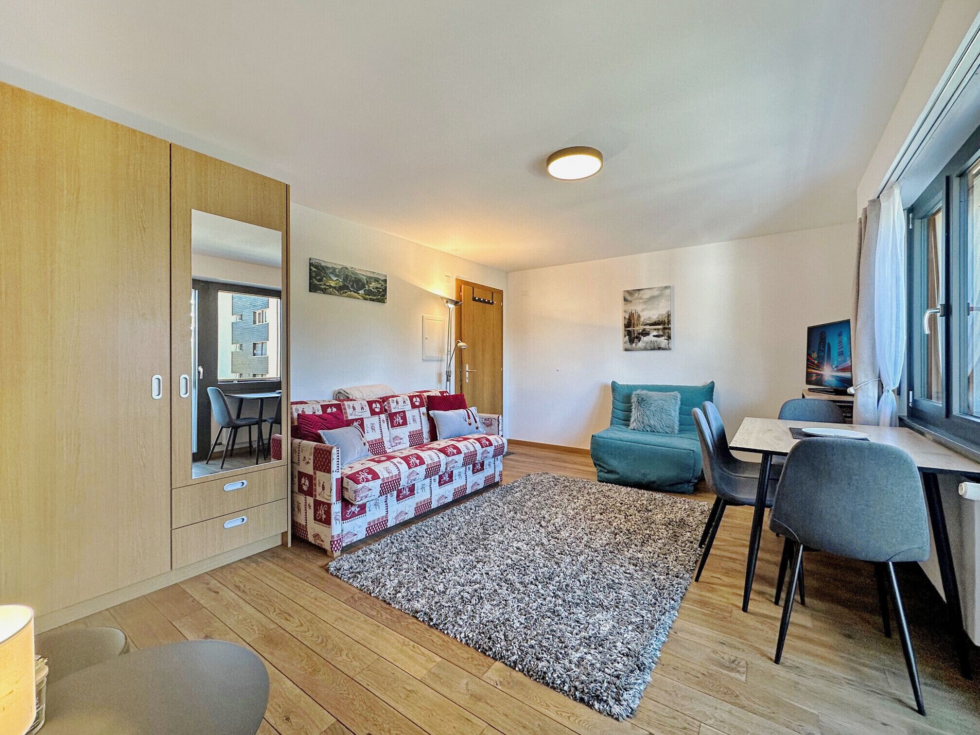 Una sala de estar moderna con un armario de madera, un sofá estampado en rojo y blanco, un sillón color turquesa, una mesa de comedor con sillas y un ventanal con cortinas.