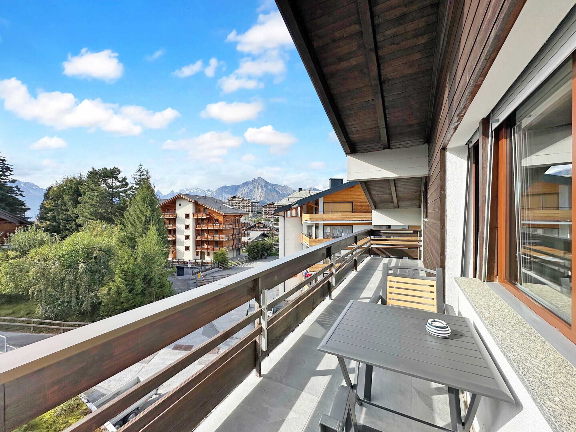 Uma varanda com mesa e cadeiras, com vista para uma área montanhosa e edifícios ao fundo.