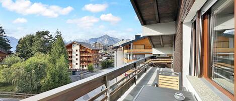 Uma varanda com mesa e cadeiras, com vista para uma área montanhosa e edifícios ao fundo.