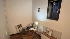 Una habitación pequeña y acogedora con suelo de baldosas, una mesa de madera con sillas, una computadora portátil, una lámpara y un pequeño banco con cojín.