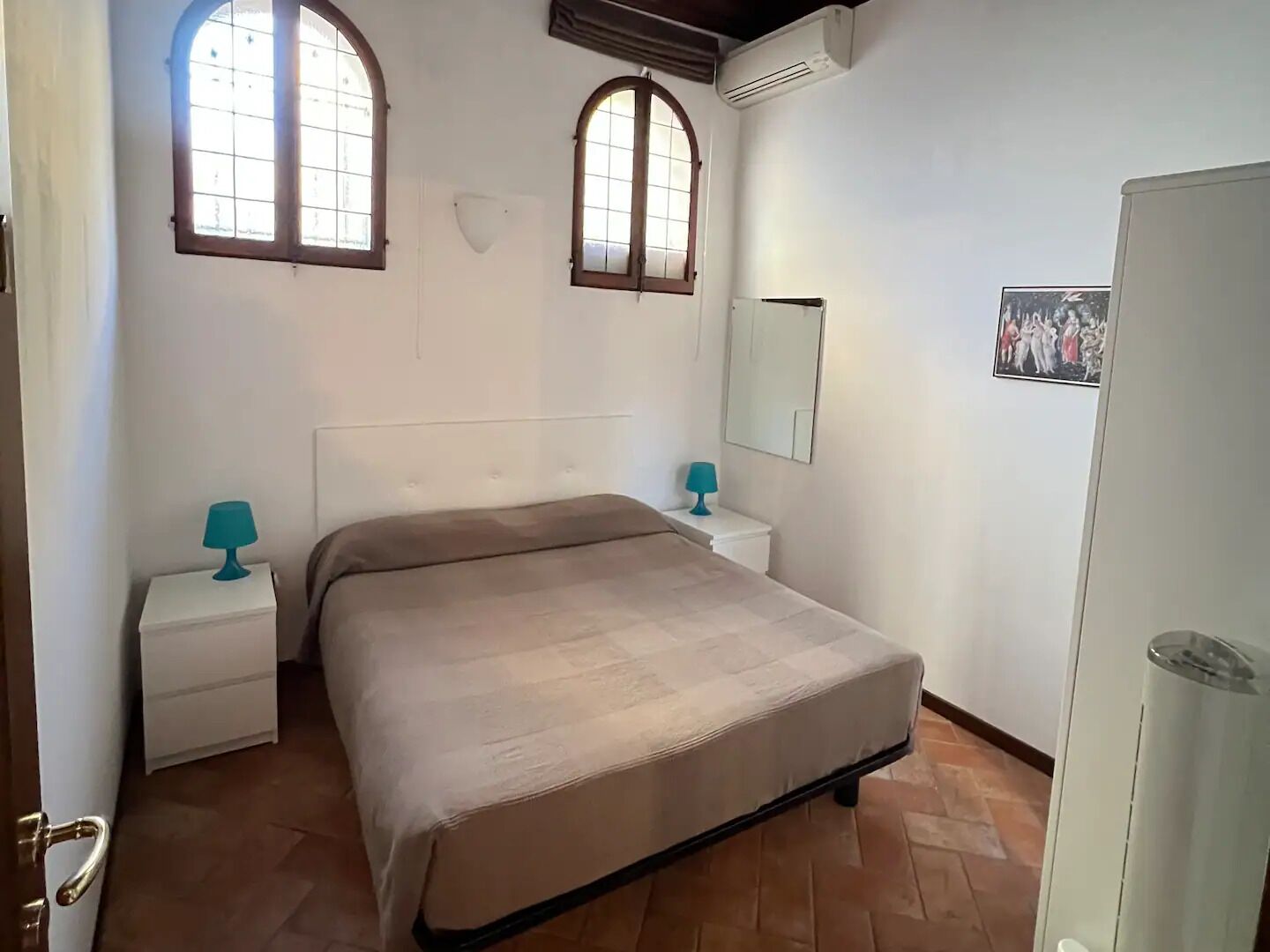 A bedroom with a bed, two bedside tables, a mirror, and a wall-mounted air conditioner.