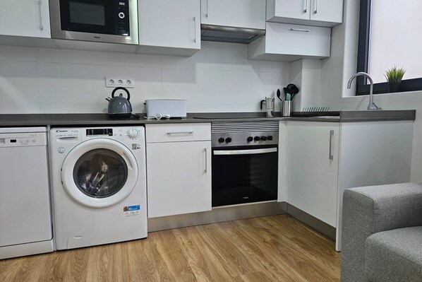 A modern kitchen with a washing machine, microwave, oven, and a grey sofa.