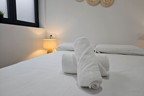 A neatly made bed with white linens and rolled towels, a bedside lamp, and a window with blinds.