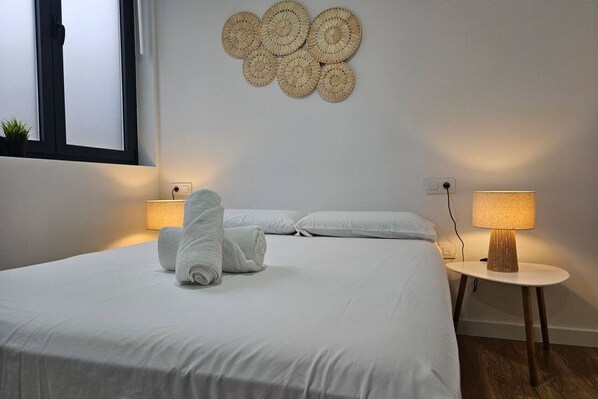 A neatly made bed with rolled towels, a wall-mounted woven artwork, a bedside lamp, and a window with a view.