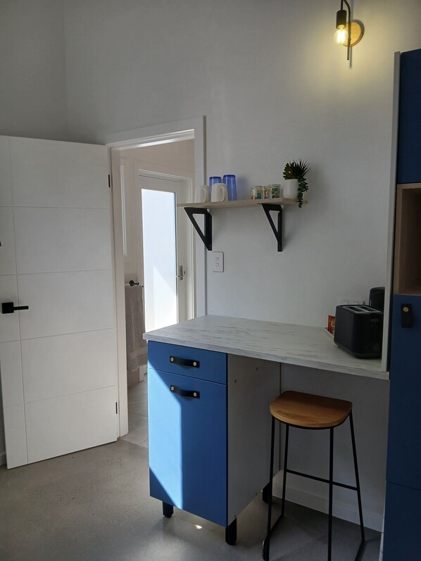 A kitchen with a white countertop, blue cabinetry, a stool, and a shelf with kitchen items.