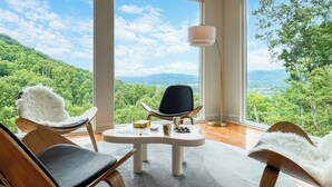 Habitación con amplias ventanas que ofrecen una vista panorámica de una cadena montañosa boscosa. Hay dos sillas de madera y una mesa con una lámpara.