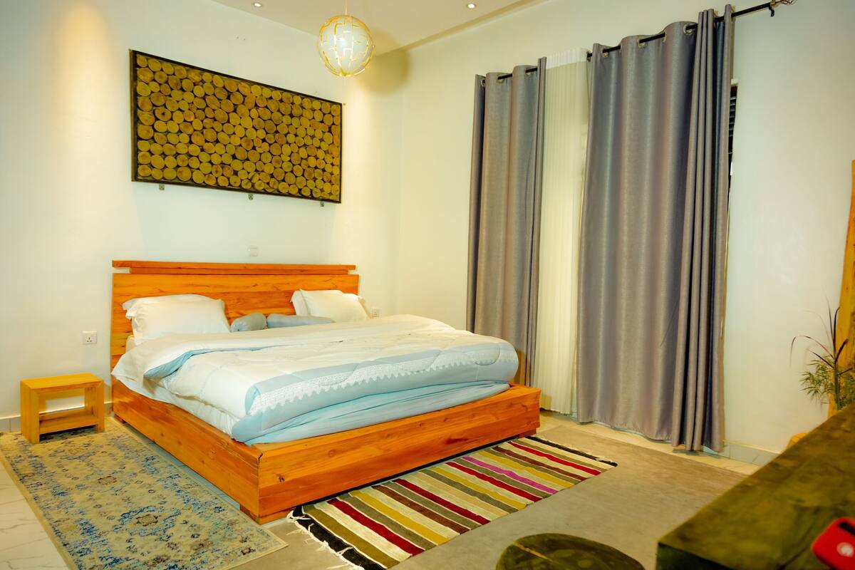 A bedroom with a wooden bed, a small bedside table, a colorful striped rug, and a wall-mounted artwork.