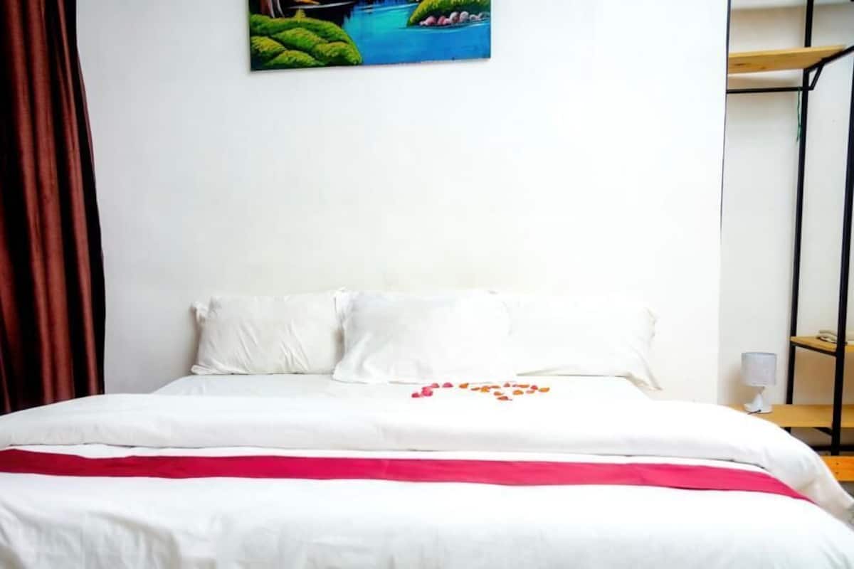 A bed with white bedding and a red blanket, a painting of a landscape on the wall, and a wooden shelf with a lamp.