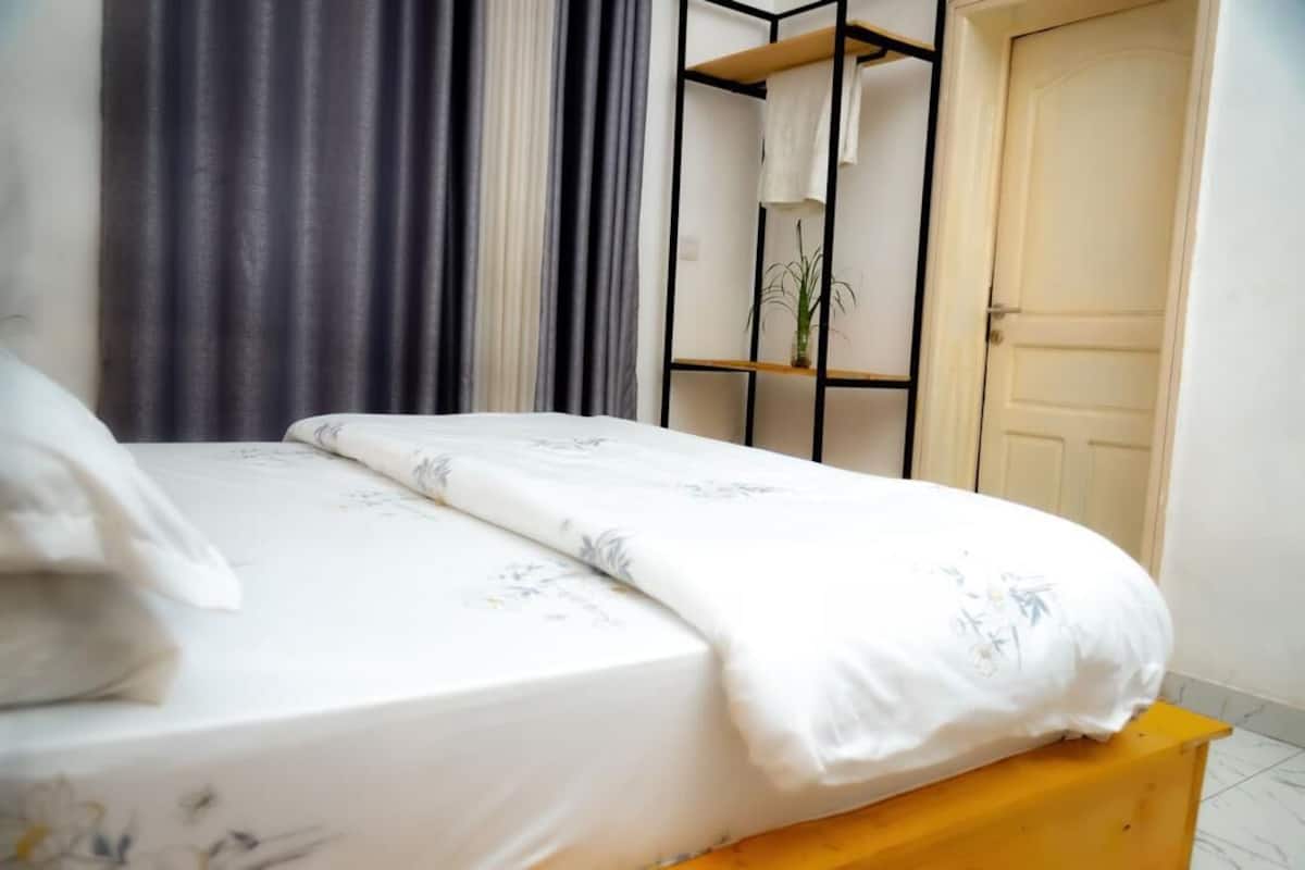 A neatly made bed with a floral bedspread and pillows, a wooden nightstand, a black metal shelf unit with a plant, and a closed door.