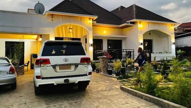 A white SUV parked in front of a house with a paved driveway and a garden area with various plants. - Esspasso  (Musanze)