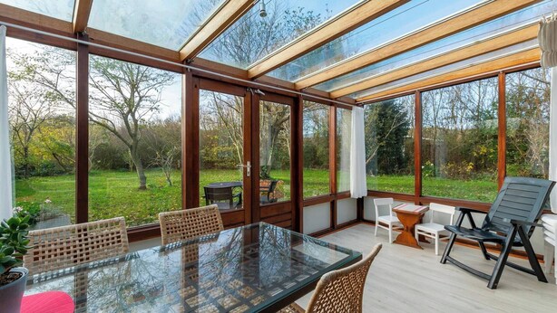 Ein Wintergarten mit Glastisch und Stühlen, umgeben von Holzrahmen und Blick auf einen Garten.