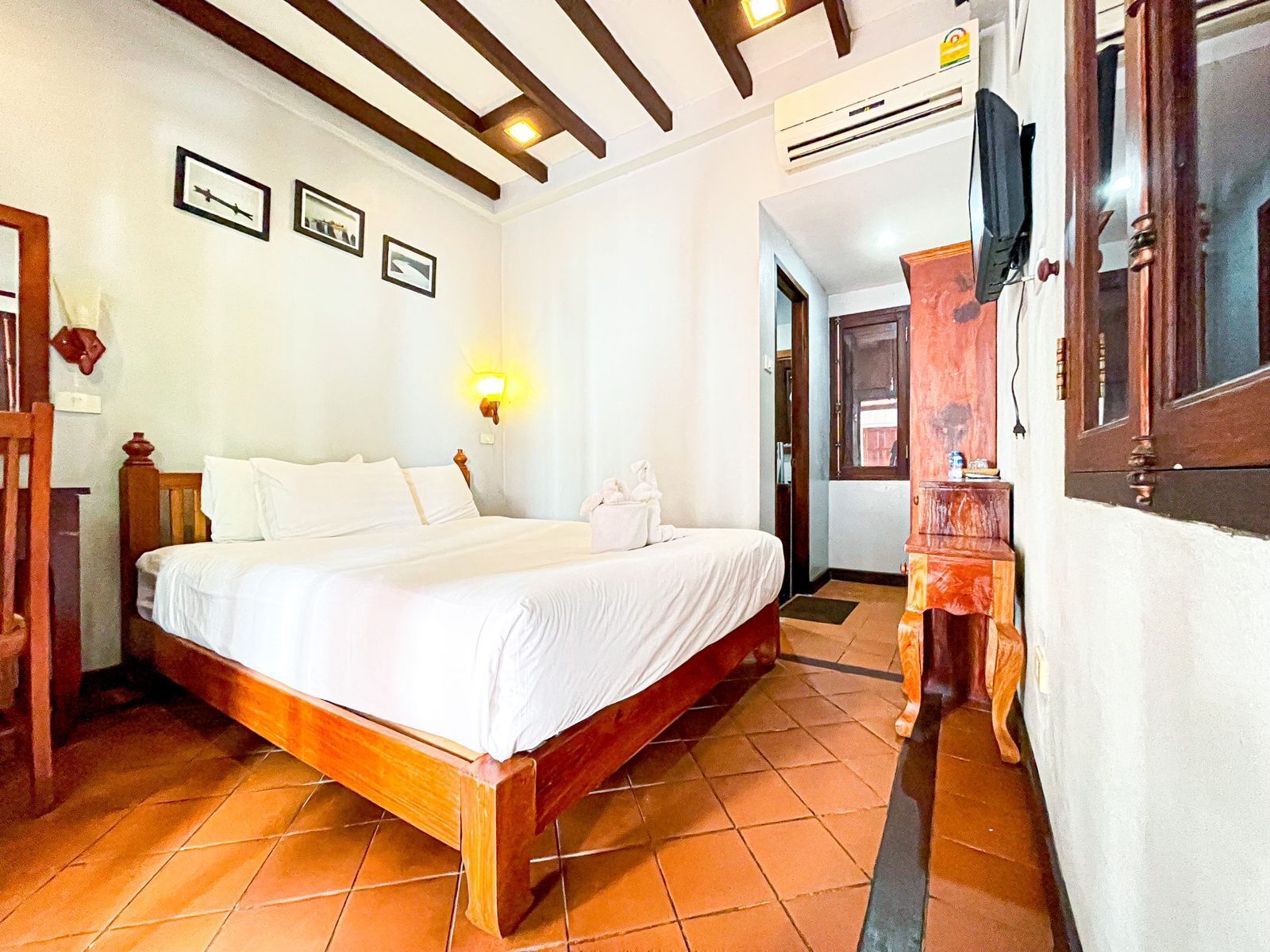 A hotel room with a wooden bed, white linens, a wall-mounted light, and a tiled floor.