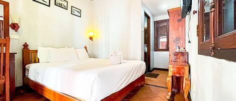 A hotel room with a wooden bed, white linens, a wall-mounted light, and a tiled floor.