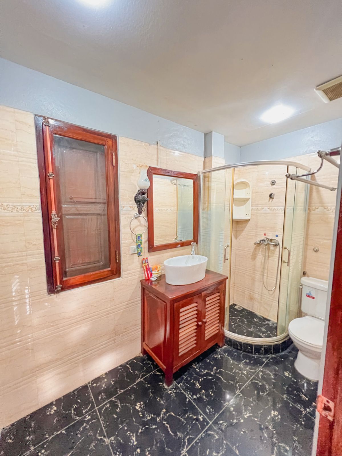 Un baño con un mueble de madera, un lavamanos blanco, un espejo y una zona de ducha con mampara de vidrio.