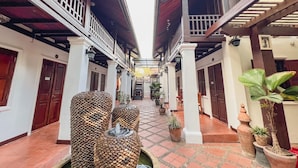 A corridor with a tiled floor, white walls, and wooden balconies. There are potted plants and a fountain along the path.