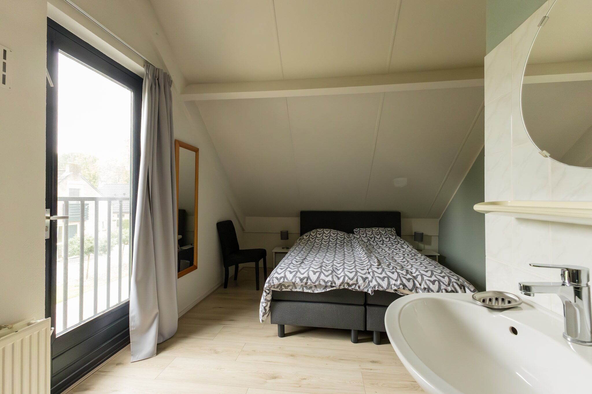 Ein Schlafzimmer mit Bett, Schreibtisch, Stuhl, einem Fenster mit Vorhängen, einem Badezimmer mit Waschbecken und einem Spiegel.