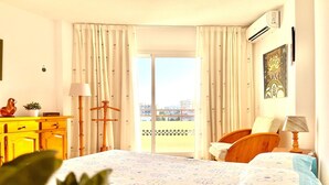 Ein Hotelzimmer mit einem Bett, einem Holzsessel, einem Schreibtisch mit Lampe, einer Pflanze und Blick auf die Stadt durch das Fenster.