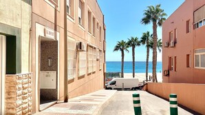 Een smalle straat met een gebouw links en een uitzicht op het strand rechts. Er staan palmbomen en een witte busje langs de straat.