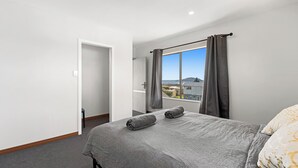 Ett hotellrum med en stor säng, grå sängkläder, utsikt över staden genom ett fönster med gardiner och en dörr som leder till ett annat rum.