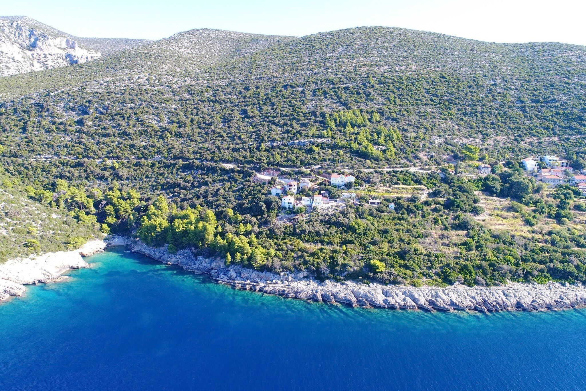 An aerial view of a coastal village with buildings nestled among green hills and a clear blue sea.