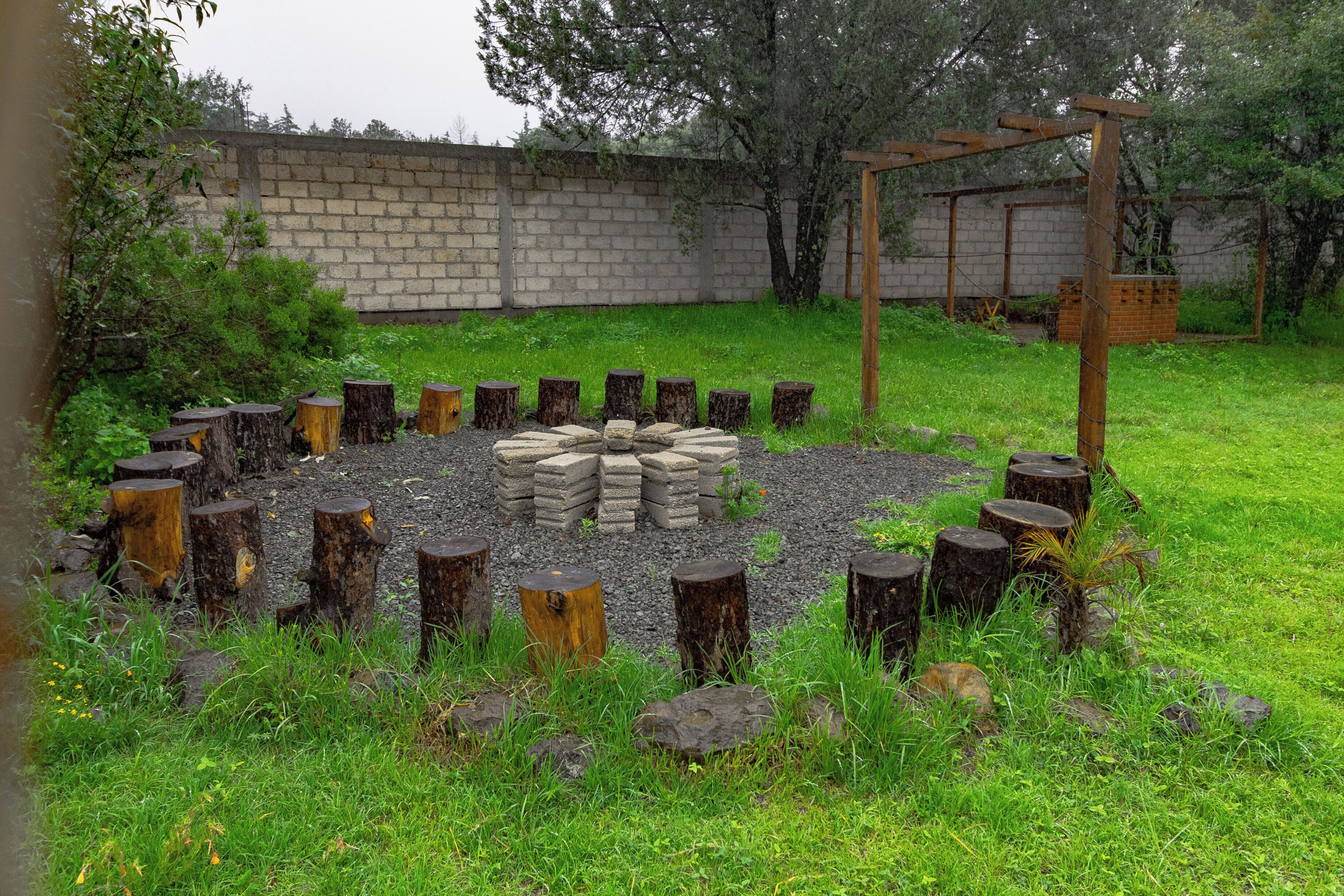 A circular arrangement of logs and stones, surrounded by grass and trees, with a wooden structure in the background.