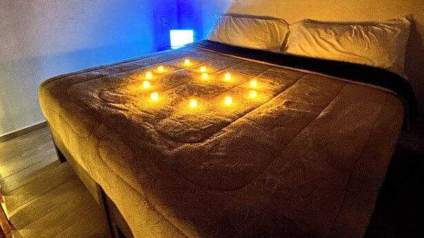 Una cama con un edredón dispuesto en forma de corazón, iluminada por velas, en una habitación con un reloj de pared y una ventana pequeña.