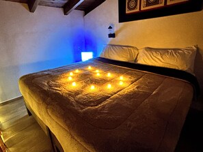 Un lit avec une couverture disposée en forme de cœur, éclairé par des bougies, dans une pièce où l’on peut voir une horloge murale et une petite fenêtre.