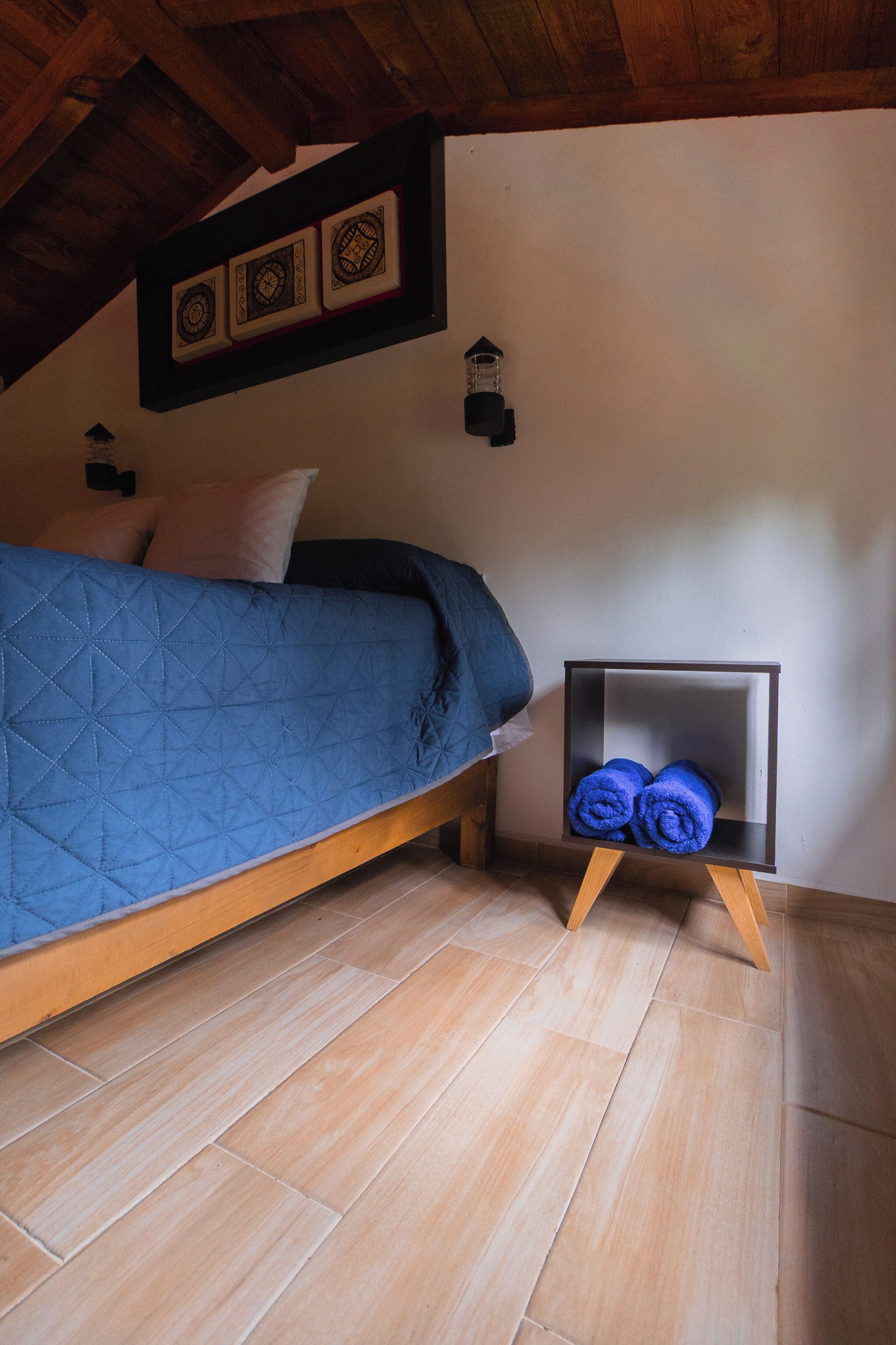 Een slaapkamer met een bed, een houten vloer, een wandplank met handdoeken en een plafond met zichtbare balken.
