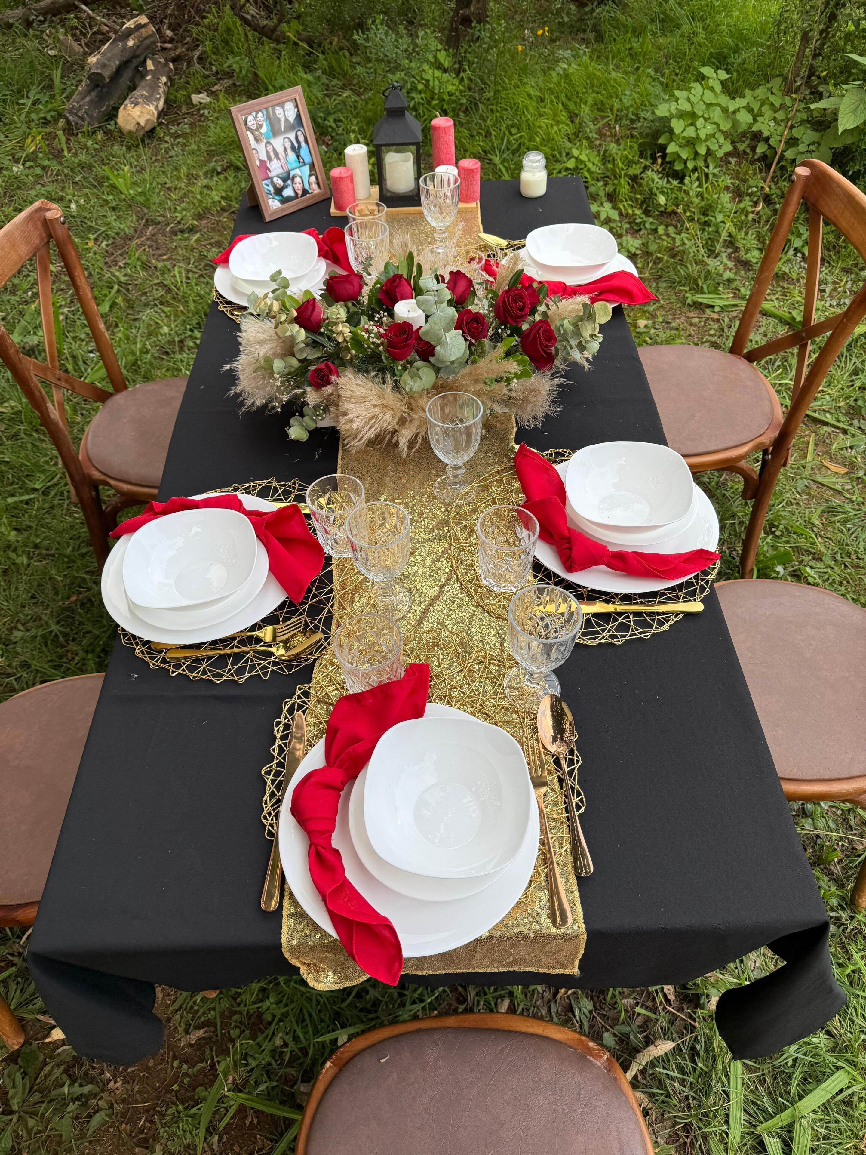 Mesa al aire libre con mantel negro, detalles dorados, servilletas rojas y un centro de mesa floral.