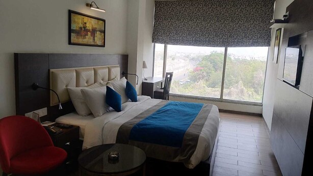 A hotel room with a bed, a small table, a chair, a desk, and a window with a view. - JB SKY HILTON INN (Lucknow)